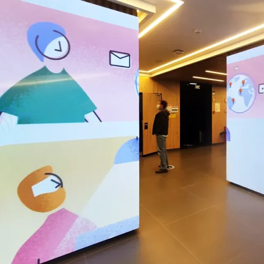 Painel LED Personalizado instalado no hall de edifício de escritórios, a revestir parede e pilar, e a reproduzir conteúdos coloridos animados, em fundo branco.