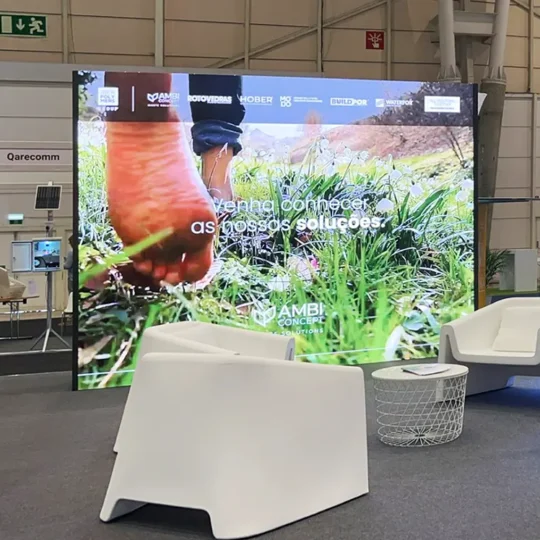 Display LED Rental em formato 4:3, a reproduzir imagem de pessoa na natureza, instalado em stand de feira, com mesa de centro e cadeiras em branco.